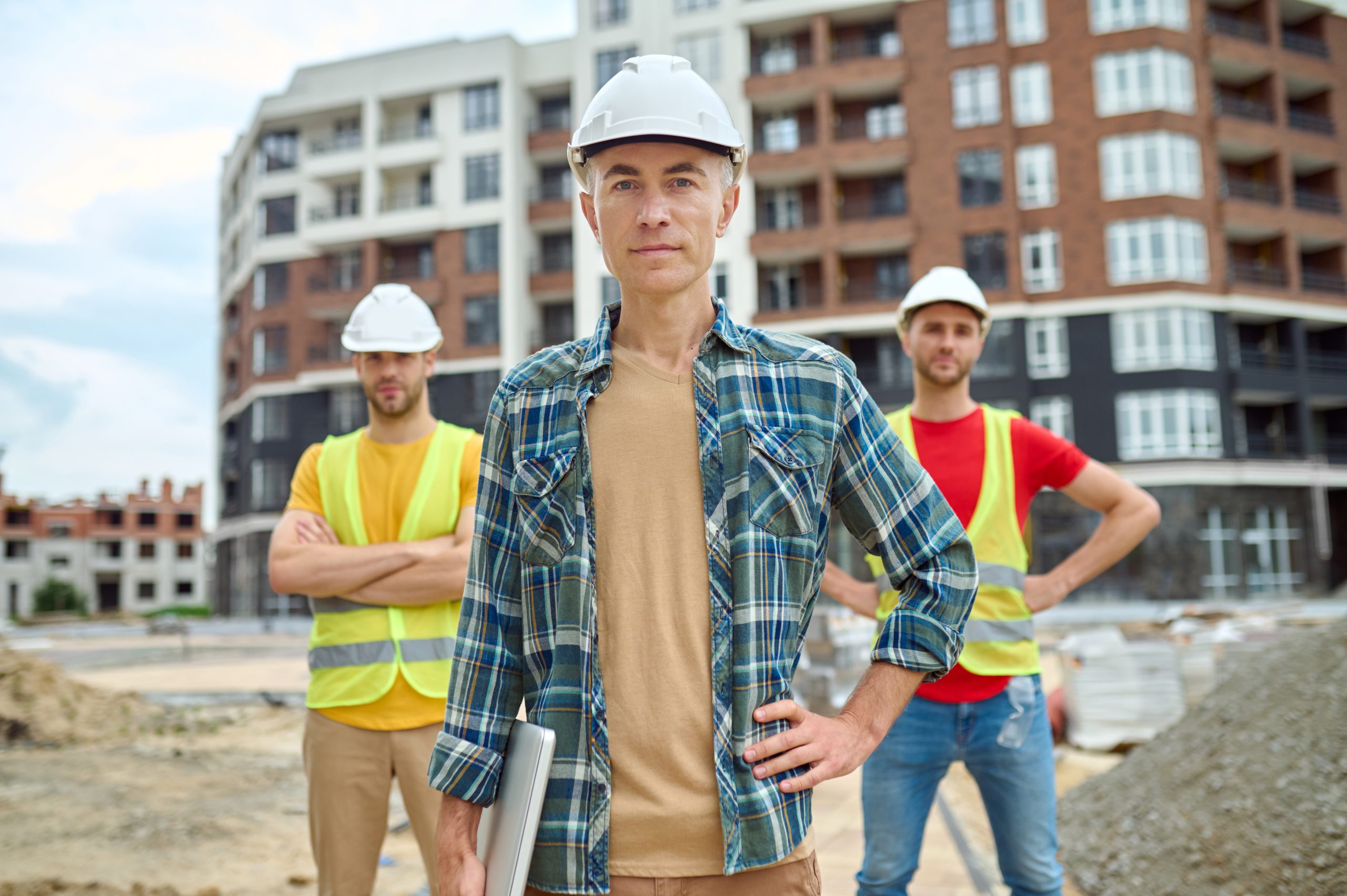 Reliability. Confident serious mature man in safety helmet and plaid shirt with laptop and two workers standing behind outdoors at construction site looking at camera